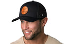 A smiling man with a trimmed beard wears the Haka Hat Golf Trucker Hat Leather | Black Club X Snapback by Haka Hat, featuring a round brown leather patch with crossed golf clubs, paired with a beige T-shirt against a white background.