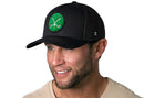 A man with light eyes and a trimmed beard smiles while wearing the Haka Hat Golf Trucker Hat Chenille | Black Club X Snapback by Haka Hat, featuring a green chenille patch with crossed golf clubs. He has on a beige shirt and stands against a white background.