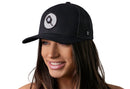 A woman with long brown hair smiles in a Haka Hat Pickleball Trucker Hat—Black Ball And Paddle Snapback by Haka Hat, featuring an embroidered black paddle and ball patch. The cap stands out against the white background.