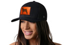 A smiling woman wears the Haka Hat Pug Trucker Hat—a black dog snapback by Haka Hat featuring a leather patch with an orange dog silhouette. The background is plain white.