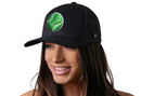 A smiling woman with long brown hair wears the Haka Hat Tennis Trucker Hat—a black mesh snapback by Haka Hat featuring a bright green embroidered tennis ball and comfort-fit sweatband. The background is plain white.
