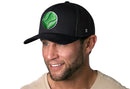 A man smiles while wearing the Haka Hat Tennis Trucker Hat, a black snapback by Haka Hat featuring a green tennis ball embroidery and comfort-fit sweatband, paired with his beige shirt against a white background.