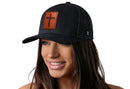 A woman with long brown hair smiles while wearing the Haka Hat Cross Trucker Hat Rectangle Leather in black, featuring a brown cross patch and adjustable snapback by Haka Hat. The background is white.