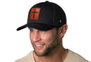 A man in a light shirt smiles while wearing the Haka Hat Cross Trucker Hat—a black mesh snapback with a rectangle brown leather patch featuring a black cross, by Haka Hat—on a plain white background.