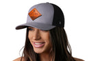 A smiling woman with long brown hair wears the Haka Hat Diamond Mountain and Trees Trucker Hat Leather, a gray-black outdoors snapback featuring a leather mountain patch, against a plain white background.