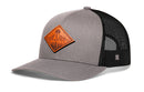 The Haka Hat Diamond Mountain and Trees Trucker Hat features a gray-black mesh snapback, curved brim, and an orange leather patch with a mountain and sun design on the front—perfect for outdoor enthusiasts.
