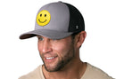 A man wearing the Haka Hat Smiley Face Trucker Hat in gray-black with a yellow emoji patch and comfort-fit snapback smiles to the side. He has light facial hair and wears a beige shirt.