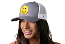 A woman smiles and looks to the side wearing the Haka Hat Smiley Face Trucker Hat—a gray-white emoji snapback from Haka Hat featuring an embroidered smiley face patch on the front.