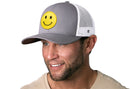A man with a trimmed beard wears the Haka Hat Smiley Face Trucker Hat, a gray-white emoji snapback from Haka Hat featuring a comfort-fit sweatband, as he smiles and looks slightly to the side.