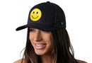 A woman with long brown hair smiles wearing the Haka Hat Smiley Face Trucker Hat, a black emoji snapback by Haka Hat with a large yellow embroidered patch. She poses against a plain white background.