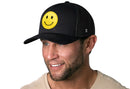 A man in a beige shirt with light stubble smiles and looks to the side, wearing the Haka Hat Smiley Face Trucker Hat—Black Emoji Snapback by Haka Hat, featuring a large yellow embroidered patch on the front.