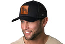 A smiling man wears the Haka Hat French Bulldog Trucker Hat Leather by Haka Hat—a black dog snapback—with a light beige shirt, against a white background.