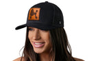 A woman smiles wearing the Haka Hat Poodle Trucker Hat—a black mesh snapback by Haka Hat featuring a brown leather patch with a poodle silhouette—set against a white background.