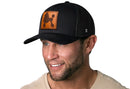 A smiling man with a short beard wears the Haka Hat Poodle Trucker Hat Leather, a black snapback featuring a brown leather patch with a poodle silhouette. He’s dressed in a light shirt against a white background.