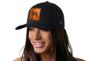 A woman with long brown hair smiles, wearing a Haka Hat Boxer Trucker Hat Leather | Black Dog Snapback by Haka Hat. The black mesh cap features a brown leather patch on the front and is set against a plain white background.