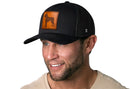 A man with a short beard and mustache smiles while looking to the side, wearing a Haka Hat Boxer Trucker Hat Leather | Black Dog Snapback by Haka Hat and a light-colored shirt.