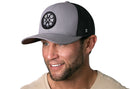 Smiling man with short brown hair and a trimmed beard wearing the Haka Hat GYM Trucker Hat in gray-black, a comfort-fit snapback with a round crossed-dumbbell logo by Haka Hat, paired with a light-colored shirt.