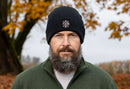 A bearded man wears a Haka Hat Pacific Northwest Beanie | Black PNW X, featuring a circular white logo, and a green zip-up jacket while standing outdoors before autumn trees with orange leaves. The background is softly blurred.