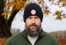 A man with a thick beard stands outdoors before autumn trees, wearing a green zip-up jacket and a black Haka Hat USA Beanie | Black USA X by Haka Hat with a woven patch.