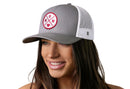 A smiling woman wears the Haka Hat Mom Trucker Hat Gray-White Snapback by Haka Hat, featuring a red embroidered MOM patch with a small heart on the front and a comfort-fit sweatband for all-day comfort.