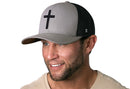 A man in a light shirt, facing right and smiling, wears the Haka Hat Cross Trucker Hat—a gray and black snapback by Haka Hat featuring a bold black cross on the front—against a white background.