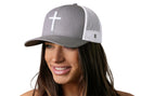 A woman with long brown hair smiles while wearing the Haka Hat Cross Trucker Hat, a gray-white snapback by Haka Hat featuring a bold white cross on the front. The background is plain white.