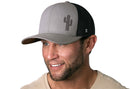A smiling man wears a beige tee and the Haka Hat Cactus Trucker Hat—Gray-Black Snapback, featuring a cactus logo and black mesh back—standing out against a white background.