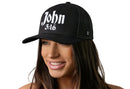 A woman smiles wearing the Haka Hat John 3:16 Trucker Hat, a black snapback by Haka Hat featuring bold white 3D puff "John 3:16" embroidery and a classic design. The background is plain white.