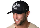A smiling man with a short beard wears the Haka Hat John 3:16 Trucker Hat, a black Christian 3D puff snapback by Haka Hat, paired with a light T-shirt against a plain white background.