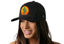 A woman wears a Haka Hat Cactus Sunset Trucker Hat—a black snapback by Haka Hat featuring a circular patch with a green cactus against an orange and yellow sunset.