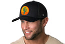 A man in a black Haka Hat Cactus Sunset Trucker Hat with a colorful cactus patch smiles and looks to the side, sporting a trimmed beard and beige shirt, radiating a relaxed western vibe against a white background.