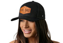 A smiling woman with long brown hair wears the Haka Hat Girl Dad Leather Trucker Hat in black, featuring an orange patch reading GIRL DAD. This snapback by Haka Hat stands out against a plain white background.