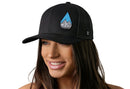 A woman with long brown hair smiles while wearing the Haka Hat Rainddrop Mountain and Trees Trucker Hat, a black outdoors snapback by Haka Hat, featuring a blue water droplet patch with mountain and trees design.