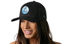 A smiling woman wears the Haka Hat Sailboat Trucker Hat— a black snapback from Haka Hat with a round patch showing a blue and white sailboat. The background is white.