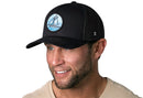 A smiling man with a trimmed beard wears a Haka Hat Sailboat Trucker Hat—a black mesh snapback by Haka Hat with a blue and white sailboat patch—and a beige shirt, set against a plain white background.