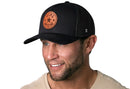 A smiling man with stubble wears the Haka Hat Girl Mom Leather Trucker Hat in black, a snapback featuring a circular GIRL MOM patch with a star center. He pairs it with a light beige shirt against a white background. Brand: Haka Hat.
