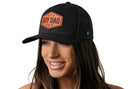 A smiling woman with long brown hair wears the Haka Hat Boy Dad Leather Trucker Hat, a black snapback by Haka Hat featuring a bold "BOY DAD" patch with small stars, standing out against a white background.