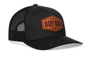 The Haka Hat Boy Dad Leather Trucker Hat by Haka Hat is a black mesh snapback featuring a brown hexagonal patch on the front that reads BOY DAD in bold letters, surrounded by five small stars.