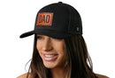A smiling woman with long brown hair wears the Haka Hat Dad Leather Trucker Hat, a black mesh snapback by Haka Hat with a brown “DAD” patch on the front, against a white background.