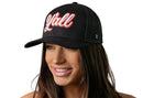 A smiling woman with long brown hair wears the Haka Hat Y'all Trucker Hat, a black script snapback by Haka Hat, featuring "Y'all" in red and white script on the front. The background is white, emphasizing its playful western style.