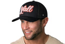 A smiling man with a trimmed beard wears the Haka Hat Y'all Trucker Hat, a black mesh snapback with red and white script. He is dressed in a light-colored shirt against a white background.