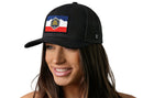 A woman with long brown hair smiles while wearing the Haka Hat Utah Flag Trucker Hat, a black snapback from Haka Hat featuring a colorful patch with a mountain, beehive, and red stripe design. The background is white.