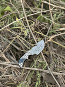 The Texan Knives Exquisite Custom Handmade Damascus Steel Guthook Skinning Knife, featuring a unique resin handle and 7.5” blade with leather sheath, rests on dry twigs and grass outdoors.