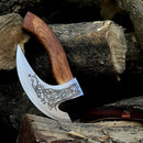 The Custom Hand-Engraved Axe Pizza Cutter by Texan Knives features a curved 8" J2 steel blade, a wooden handle with intricate patterns, and comes with a leather sheath, displayed beside stacked logs.