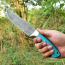 A hand holds the Texan Knives Custom Handmade Damascus Skinning Knife – 4.5" Blade, featuring a patterned steel blade and a turquoise handle with gold accents, set against lush green foliage and trees outdoors.