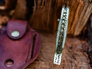 Close-up of the Texan Knives Custom Handmade Fish Style Damascus Steel Folding Knife with sheep horn & brass handle, shown upright on wood beside its included brown leather sheath.