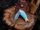 The Texan Knives Custom Handmade Fish Style Damascus Steel Folding Knife features an epoxy resin handle with brass accents and comes with a leather sheath, displayed on a cut tree stump.