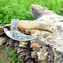 The Custom Hand Engraved Ax Pizza Cutter with J2 Steel Blade by Texan Knives features a carved wooden handle and rests on a weathered tree trunk, showcasing craftsmanship and utility against lush green foliage in the background.