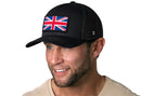 A smiling man with a short beard wears the Haka Hat United Kingdom Flag Trucker Hat, a black Union Jack snapback by Haka Hat, paired with a light shirt and standing against a plain white background.