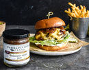 A quart jar of Terrapin Ridge Farms Balsamic Onion Jam sits by a cheeseburger with lettuce on a bun, ideal for boosting burgers or charcuterie boards, with fries in a small metal bucket on a gray tabletop.